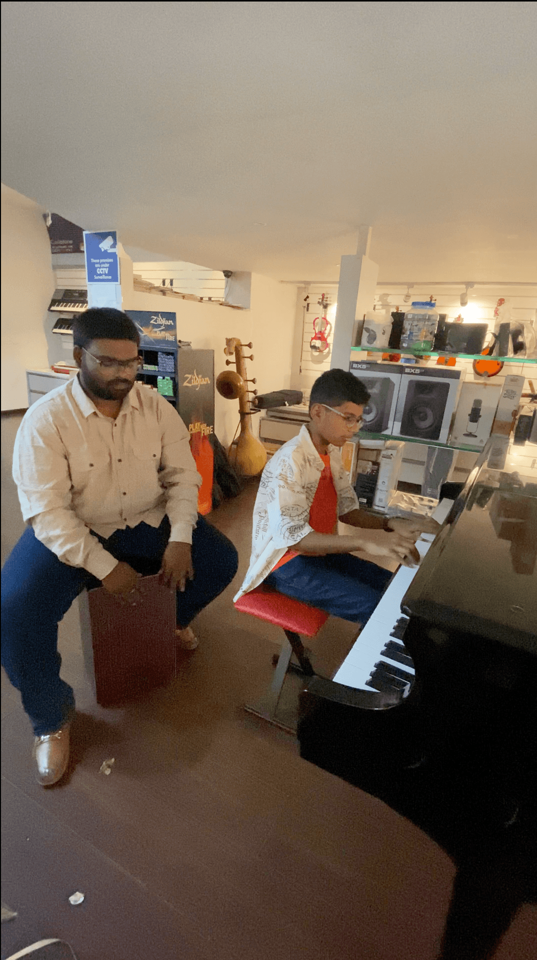Bala playing a black piano while another musician sits beside him holding a cajon.