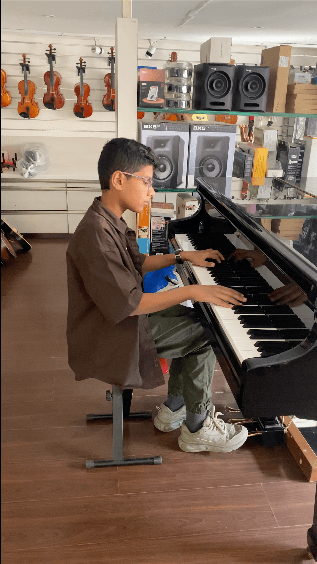 Bala seated in profile at a black piano, both hands on the keys, with string instruments on the wall behind him.