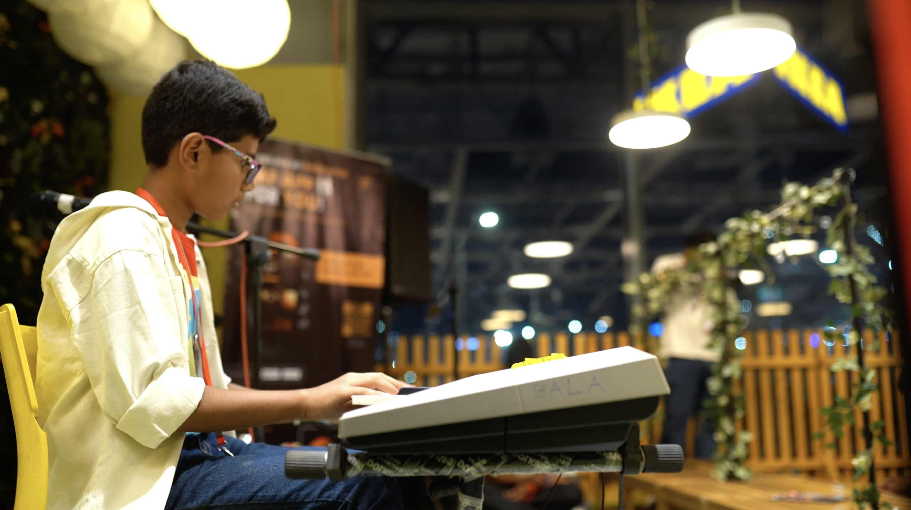 Bala seated at a keyboard on a yellow chair, with an IKEA sign and stage lights behind him.