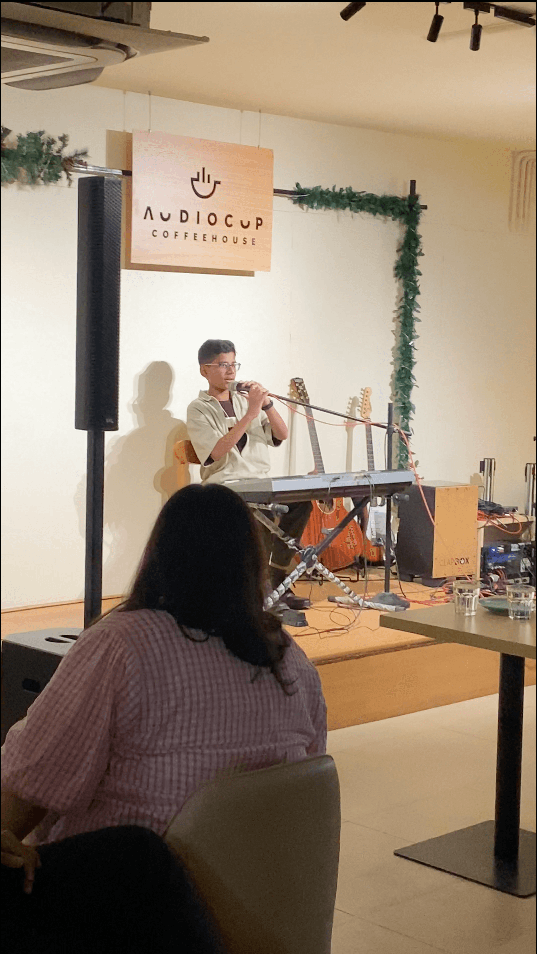 Bala on a small stage at Audiocup Coffeehouse, playing a melodica, with a keyboard in front of him and a listener in the foreground.