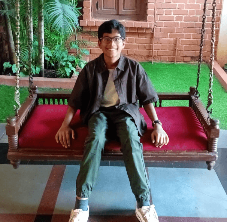 Bala seated on a wooden swing with a red cushion, smiling, in front of a brick wall and plants.